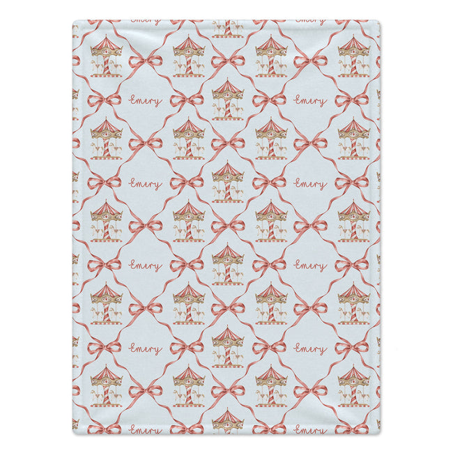 A rectangular fabric item, likely a blanket or towel, featuring a repeating pattern of carousel illustrations, red bows, and the name 'Emery' in cursive on a pale blue background.