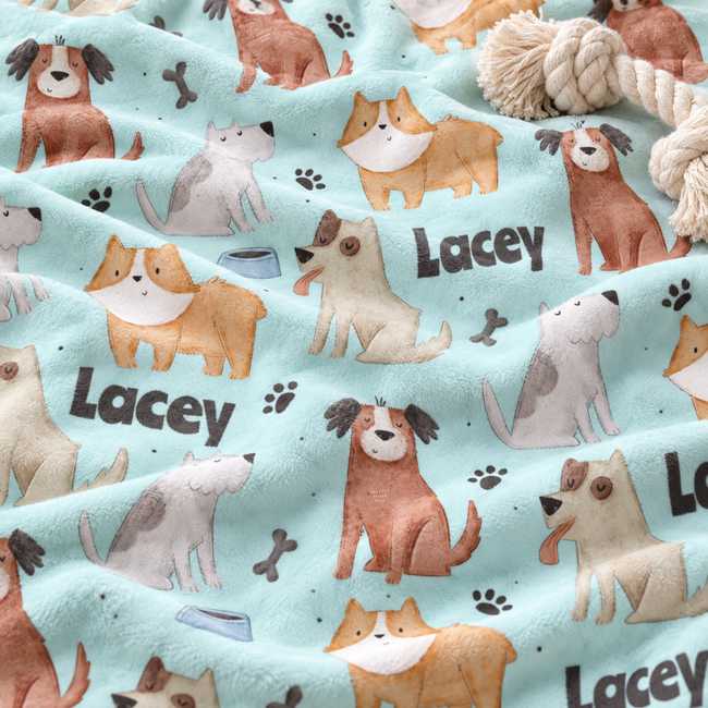 A close-up of a soft fleece blanket with a light blue background, printed with various cartoon dog illustrations, paw prints, bones, and the name 'Lacey' in dark lettering. A chunky cream rope toy is draped on top of the blanket.