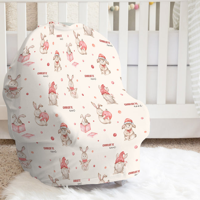 A baby car seat cover with a festive Christmas bunny print, styled on a white faux fur rug in front of a white wooden crib.