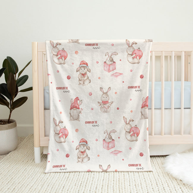 A baby blanket with a cute watercolor bunny pattern is draped over a light wood crib in a nursery setting. The blanket features white rabbits wearing pink and red Santa hats and holding pink gift boxes, with the name 'CHARLOTTE' and 'love' printed in pink.