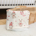 A folded cream-colored baby blanket with a cute watercolor print of rabbits, gnomes, and pink polka dots, resting on a white fluffy rug next to a woven straw basket.