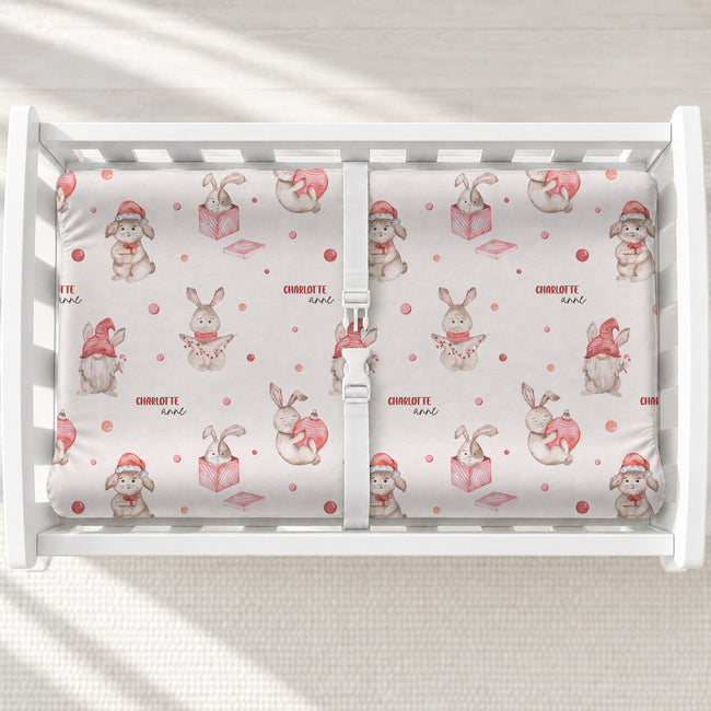 A white changing table with a personalized changing pad cover featuring a repeating pattern of cute cartoon bunnies in pink and red, with the text 'CHARLOTTE Anne' printed on the fabric.