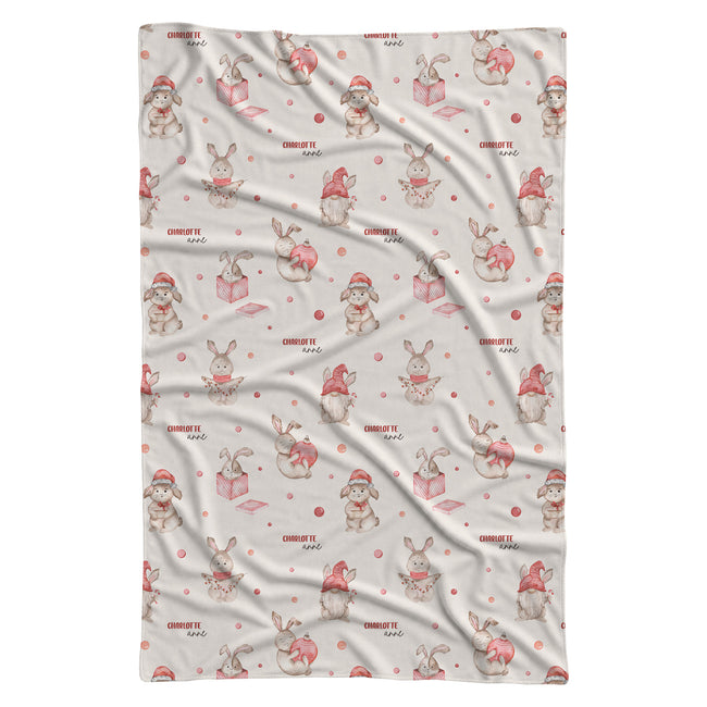 A personalized swaddle blanket with a repeating Christmas bunny pattern, featuring light brown rabbits in red Santa hats next to pink gift boxes on a warm off-white background.