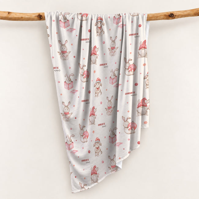 Personalized Christmas swaddle blanket hanging on a wooden rail with watercolor bunnies, holiday details and custom name Charlotte Anne.