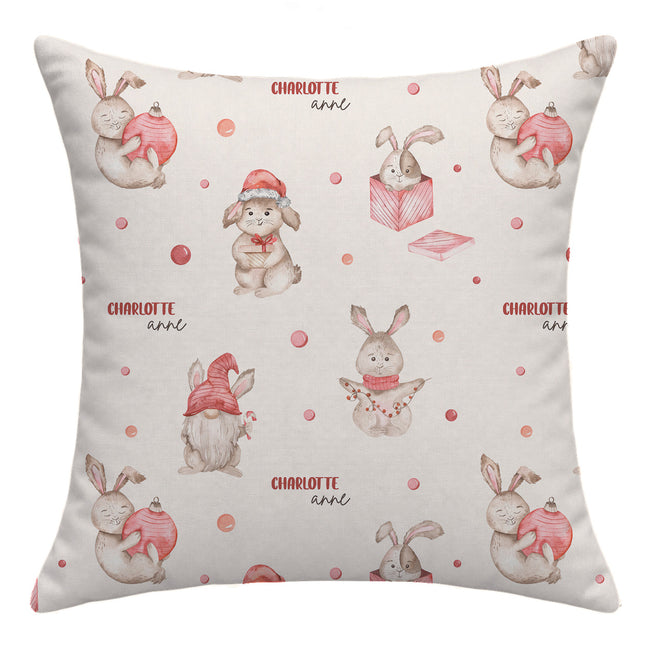 A square throw pillow featuring a repeating watercolor pattern of rabbits in winter clothing, including Santa hats and scarves, holding pink ornaments, with the name 'CHARLOTTE' and 'ann' printed in red text. The background of the pillow is a warm cream color.