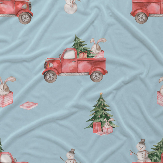 Light blue fabric with a repeating watercolor-style holiday print featuring red pickup trucks carrying Christmas trees, snowmen, rabbits, and wrapped presents.
