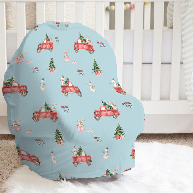 A baby car seat cover with a light blue background and a festive Christmas print featuring red pickup trucks, Christmas trees, snowmen, and the words 'Santa's toys', displayed on a white fluffy rug in front of a white crib.