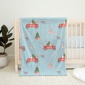 A light blue baby blanket with a festive Christmas print featuring red pickup trucks, snowman, rabbits, and Christmas trees, draped over a white wooden baby crib.