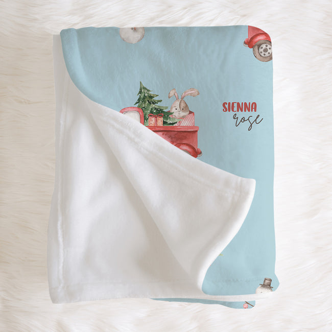 A personalized baby blanket with a light blue printed side featuring a red truck, bunny, tree, and the name 'SIENNA rose', paired with a soft white sherpa reverse, displayed on a white faux fur background.