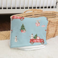 A light blue baby blanket with a Christmas-themed print featuring red trucks, a snowman, a rabbit, and a Christmas tree, draped over a woven straw basket.