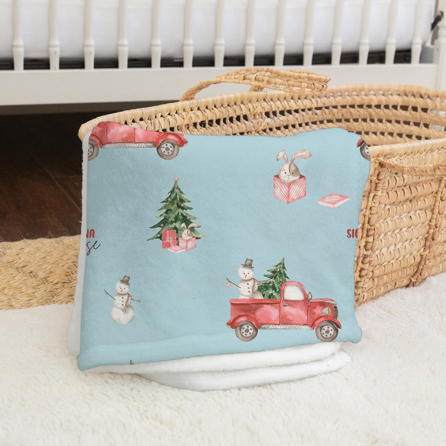 A light blue baby blanket with a Christmas-themed print featuring red trucks, a snowman, a rabbit, and a Christmas tree, draped over a woven straw basket.