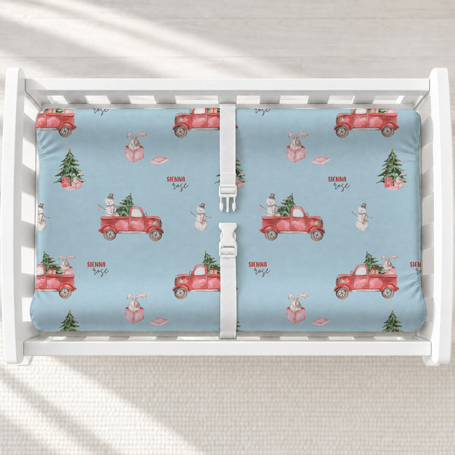 A white changing table with a personalized changing pad cover featuring light blue background and a festive Christmas theme, featuring red pickup trucks carrying Christmas trees and gifts, snowmen, and small trees. 