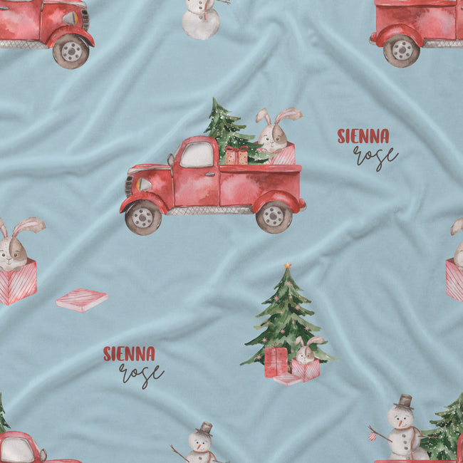 A light blue fabric with a festive Christmas print featuring red vintage pickup trucks carrying Christmas trees, snowmen, bunnies, and gift boxes. The text 'SIENNA rose' is printed on the fabric.