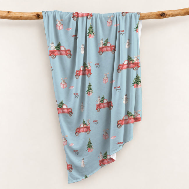 Personalized Christmas swaddle blanket hanging on a wooden rail with red vintage trucks, Christmas trees, snowmen, bunnies and custom name.