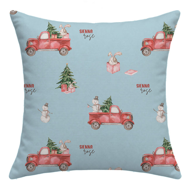 A square throw pillow with a light blue background featuring a repeating watercolor pattern of red pickup trucks carrying Christmas trees, snowmen, rabbits in gift boxes, and the text 'SIENNA rose'.