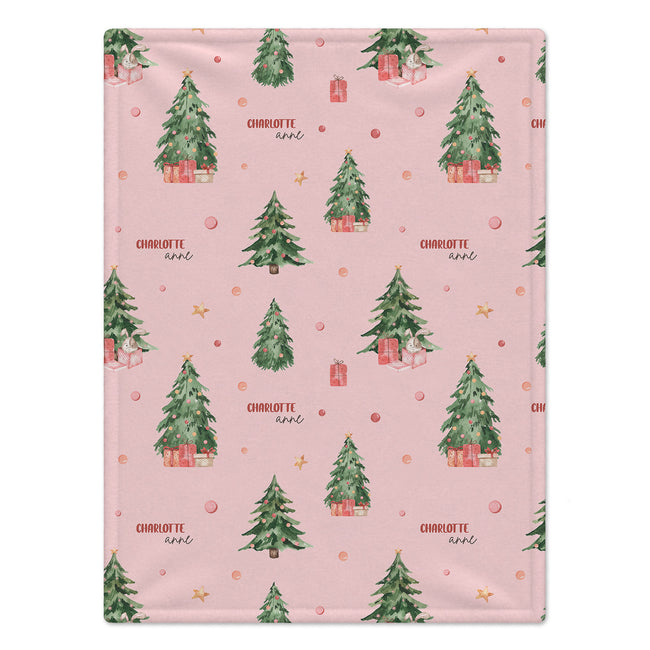 A personalized blanket featuring a repeating Christmas pattern of green watercolor trees, wrapped gifts, and gold stars on a soft pink background. The word 'CHARLOTTE' is printed in a cursive font next to the trees.