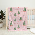 A pink baby blanket with a Christmas tree pattern draped over a white wooden crib in a nursery setting.