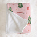 A folded baby blanket with a pink Christmas print and a white sherpa fleece backing, displayed on a white faux fur background. The blanket features watercolor-style green Christmas trees, red ornaments, wrapped gifts, and the personalized name 'Charlotte ann' printed in black script.