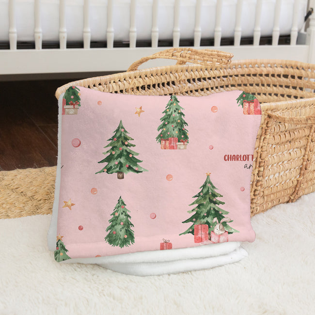 A pink baby blanket with a Christmas tree pattern and the name 'CHARLOTTE' printed on it, styled in a nursery setting next to a woven basket.