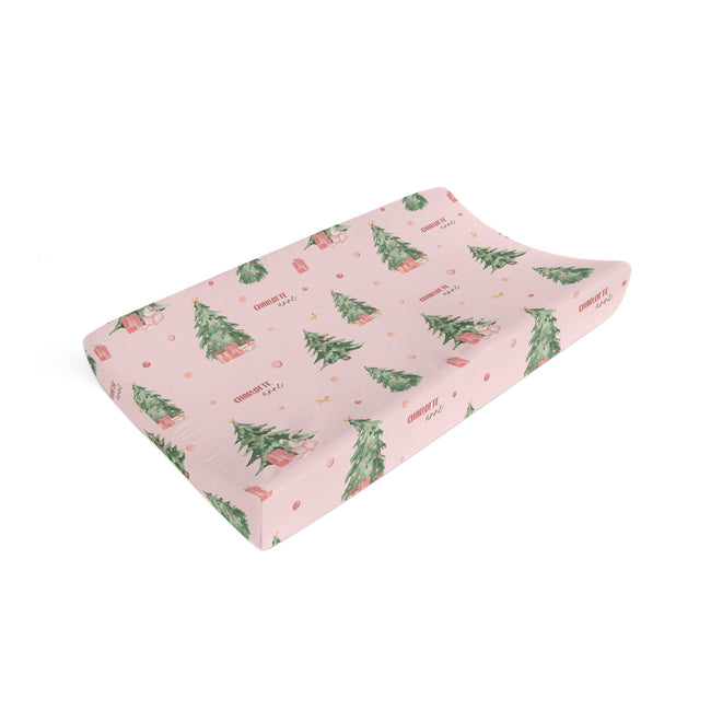A pink changing pad cover featuring a watercolor Christmas tree and cabin pattern, shown on a clean white background.