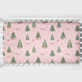 A crib mattress fitted sheet with a festive Christmas pattern featuring green pine trees, small red gift boxes, and gold stars on a light pink background. The sheet is shown fitted inside a white wooden crib.