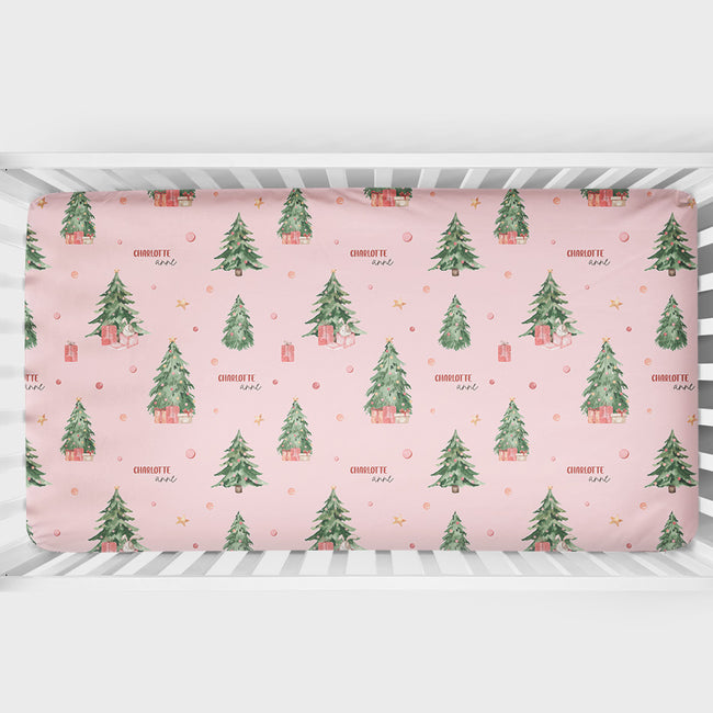 A crib mattress fitted sheet with a festive Christmas pattern featuring green pine trees, small red gift boxes, and gold stars on a light pink background. The sheet is shown fitted inside a white wooden crib.