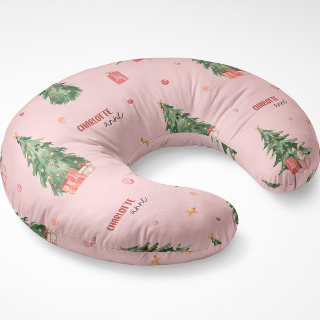 A U-shaped nursing pillow in light pink with a Christmas print featuring green trees, red gifts, and the text 'CHARLOTTE and'.