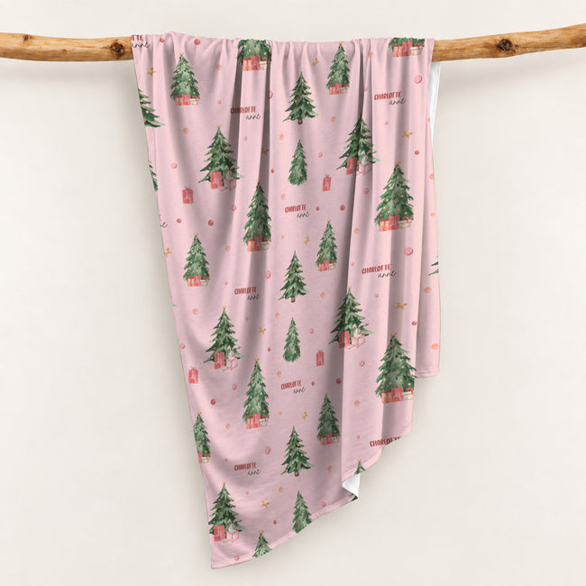 Personalized Christmas swaddle blanket hanging on a wooden rail with watercolor Christmas trees, presents and custom baby name.