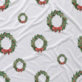White fabric with a repeating pattern of green holly wreaths decorated with red berries and red bows.