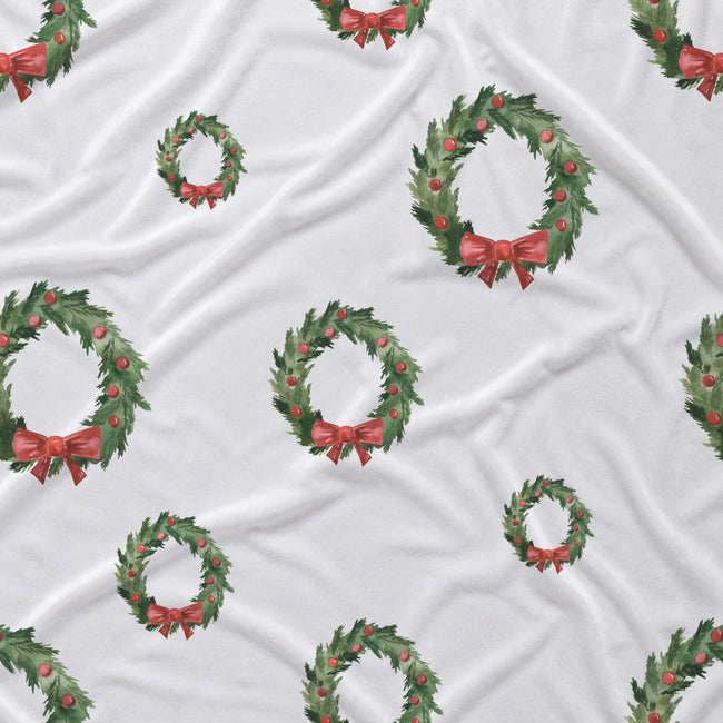 White fabric with a repeating pattern of green holly wreaths decorated with red berries and red bows.