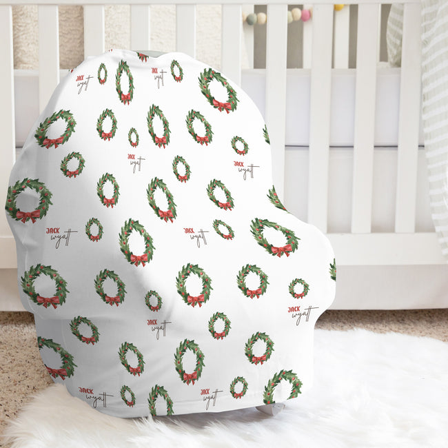 A white baby car seat cover with a festive Christmas wreath pattern, displayed on a white faux fur rug in front of a white wooden crib.
