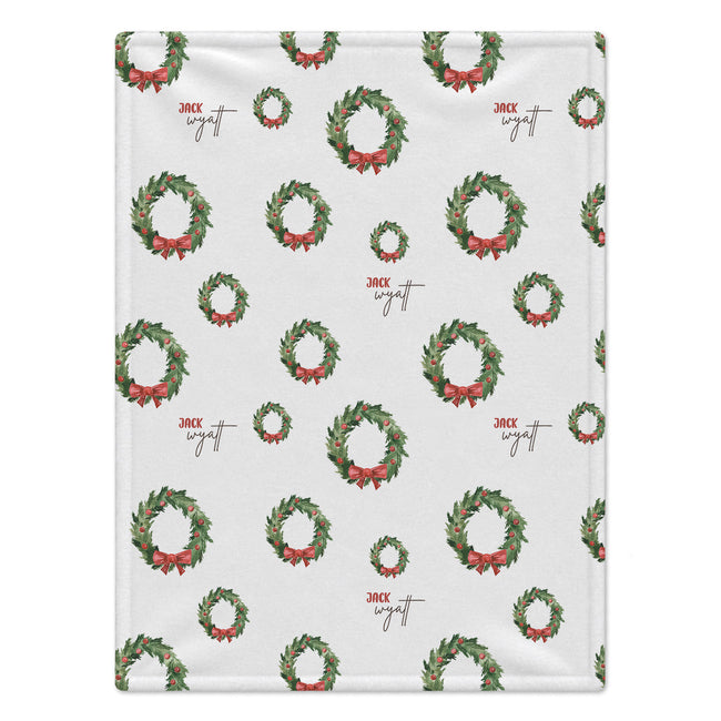 A personalized blanket featuring a repeating pattern of green Christmas wreaths with red bows and the brand name 'Jack Wyatt' printed in red and brown script.