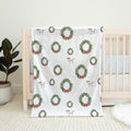 A white baby blanket with a repeating Christmas wreath and red bow pattern, draped over a light wood crib in a nursery setting.