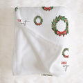 A white baby blanket with a Christmas wreath and red bow pattern, laid flat on a white faux fur background.