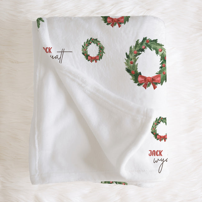 A white baby blanket with a Christmas wreath and red bow pattern, laid flat on a white faux fur background.