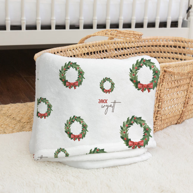 A white blanket with a green Christmas wreath pattern and red bows, folded on a white rug next to a woven basket. The blanket has a custom name printed in the center: 'JACK wyatt'.