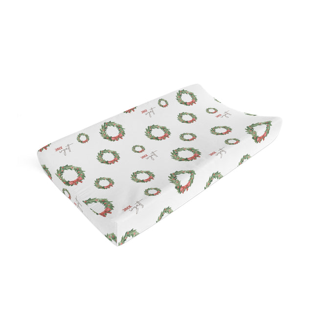 A white baby changing pad cover featuring a repeating pattern of green wreaths with red bows, shown on a clean white background.