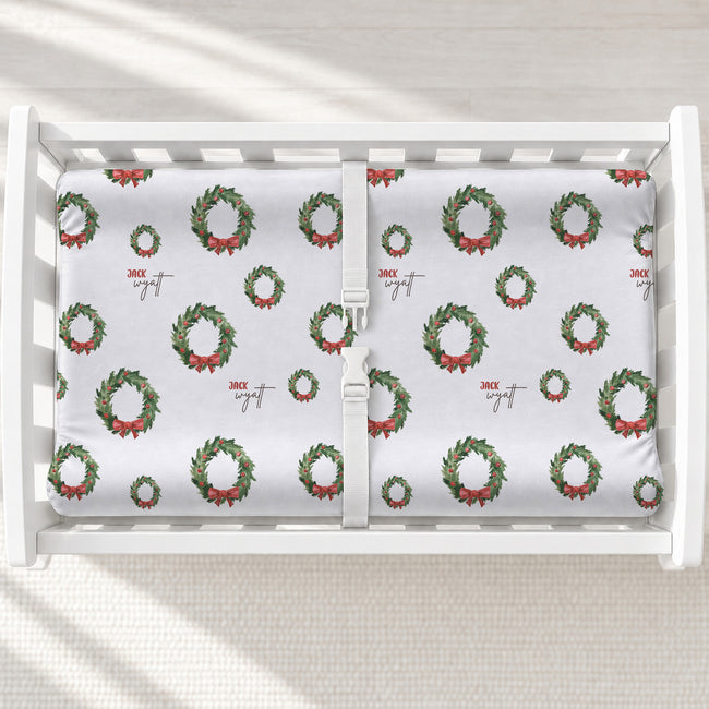 A white changing table with a personalized changing pad cover featuring white background and a green Christmas wreath pattern with red bows.
