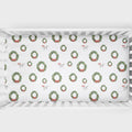 A white crib mattress fitted sheet with a repeating Christmas wreath pattern in green and red, shown inside a white crib.