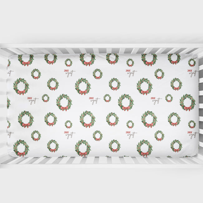 A white crib mattress fitted sheet with a repeating Christmas wreath pattern in green and red, shown inside a white crib.