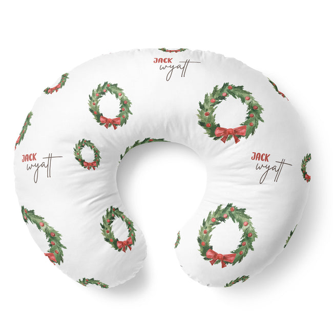A white U-shaped nursing pillow featuring a repeating pattern of green holly wreaths with red bows and the name 'JACK wyatt' printed in brown script.