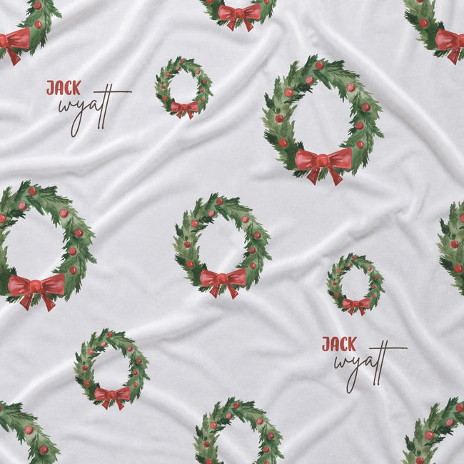 White fabric with a repeating pattern of green holly wreaths decorated with red berries and red bows, interspersed with the text 'JACK wyatt' in brown script.