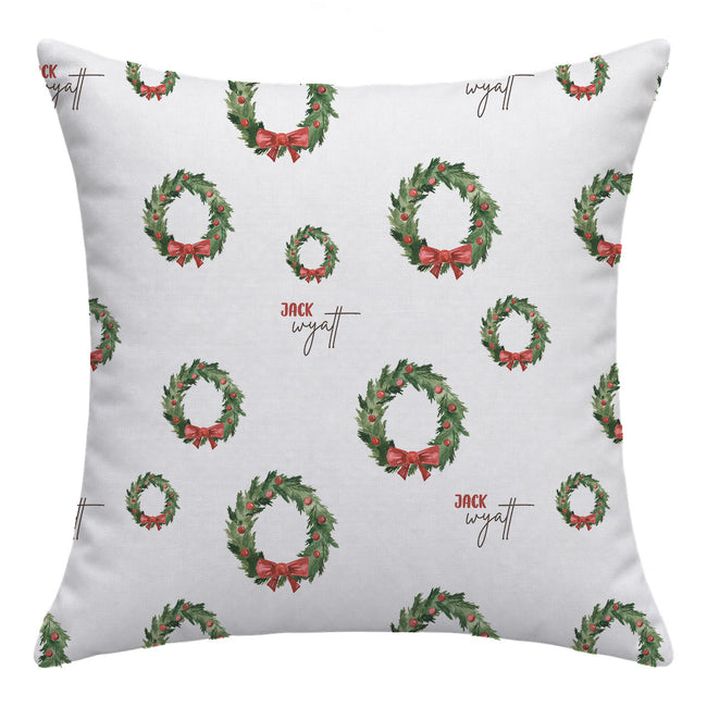 A square throw pillow with a white background featuring a repeating pattern of green Christmas wreaths with red bows and the text 'Jack Wyatt' in brown script.