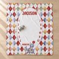 A personalized baby milestone blanket laid flat on a light wood floor. The blanket features a colorful harlequin diamond border in red, blue, yellow, green, and orange on a white background, with a large white oval center printed with the name 'JACKSON' and a month countdown from 1 to 12.