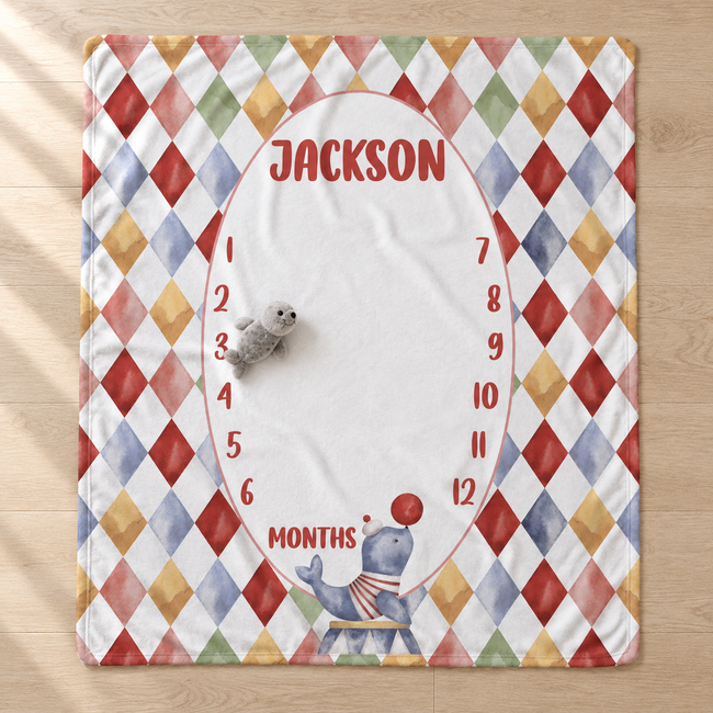 A personalized baby milestone blanket laid flat on a light wood floor. The blanket features a colorful harlequin diamond border in red, blue, yellow, green, and orange on a white background, with a large white oval center printed with the name 'JACKSON' and a month countdown from 1 to 12.