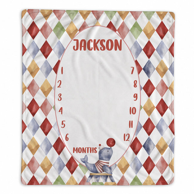 A personalized baby monthly age tracker blanket featuring a colorful harlequin diamond pattern border, a central white oval space for a photo, and red text reading 'JACKSON' and 'MONTHS' with numbers 1 through 12. A small juggling bear illustration is at the bottom.