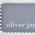 A personalized baby crib fitted sheet  in a dusty slate blue color with the names 'oliver James' printed in a cream serif font, shown inside a white wooden crib.