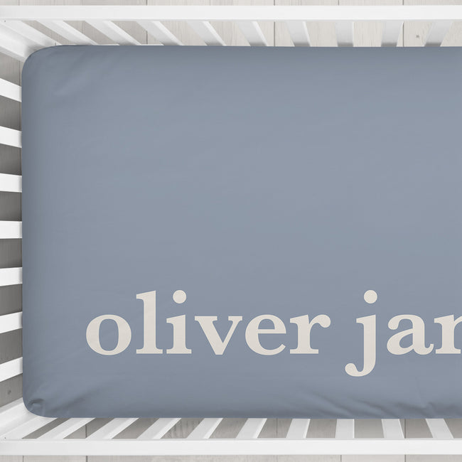 A personalized baby crib fitted sheet  in a dusty slate blue color with the names 'oliver James' printed in a cream serif font, shown inside a white wooden crib.