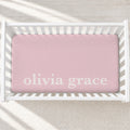 A pink fitted crib sheet with the personalized name 'olivia grace' printed in white script, shown inside a white wooden crib on a light textured rug.