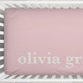 A dusty pink fitted crib sheet with the name 'olivia grace' printed in white lowercase serif letters, shown inside a white wooden crib.